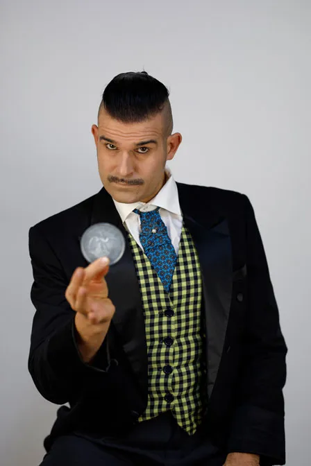 Butler Herrmann hält eine große Silbermünze in der Hand, trägt einen schwarzen Frack und eine gelbe Weste, sein Blick ist streng und seine Haare sind nach hinten gegelt