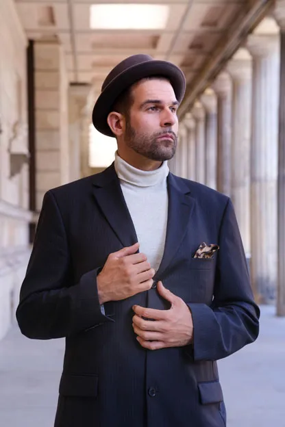 Elias Elastisch in einem dunkelblauen Nadelstreifen-Anzug, trägt einen Hut und einen weißen Rollkragen-Pullover, hat einen Drei-Tage-Bart, aufgenommen auf der Museumsinsel in Berlin