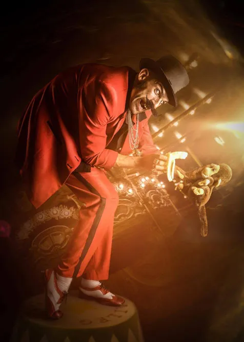 Zirkus Direktor Michal Popowize hält einen Plüschaffen im Arm und möchte ihn mit einer Banane füttern, im Hintergrund eine dunkle Zirkus-Manege, trägt Lackschuhe und eine rote Uniform