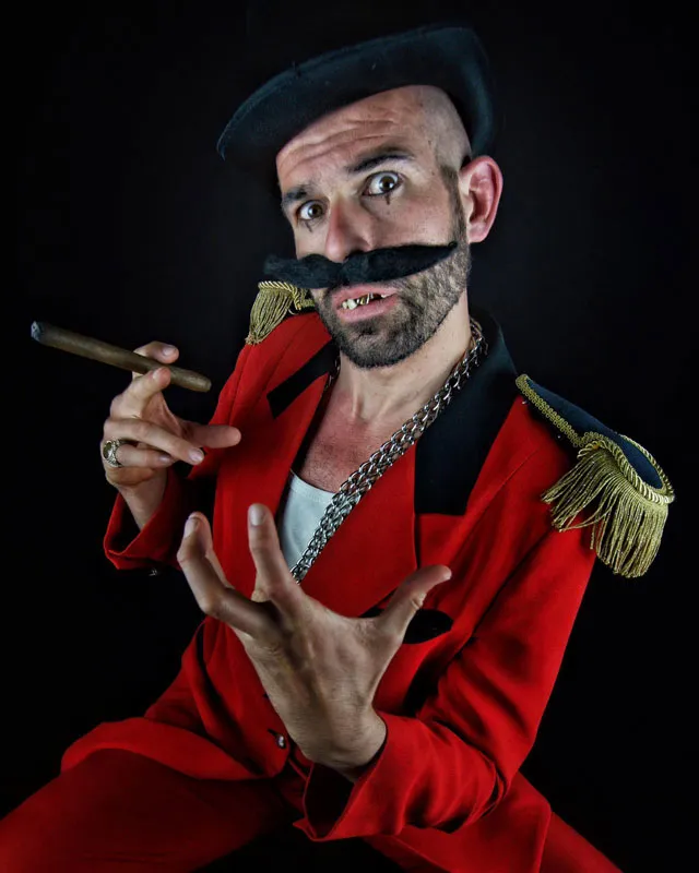 Zirkus Direktor Michal Popowize steht in einem kleinen graugrünen Raum im Morgenmantel, trägt eine runde rote Sonnenbrille und schwarze Lackschuhe mit weißen Sportsocken, im Hintergrund ein kleiner Stoffaffe im Fensterrahmen