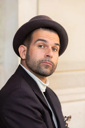 Elias Elastisch in einem dunkelblauen Nadelstreifen-Anzug, trägt einen Hut und einen weißen Rollkragen-Pullover, hat einen Drei-Tage-Bart, aufgenommen auf der Museumsinsel in Berlin