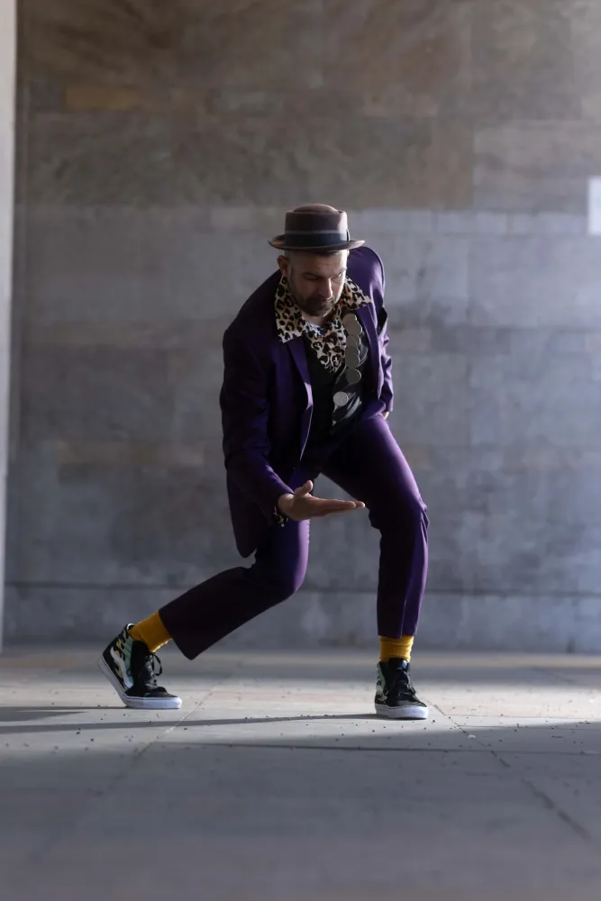 Elias Elastisch in dynamischer Tanzpose, trägt einen lila Anzug, einen Pork-Pie-Hut, Leoparden-Schal und gelbe Socken, vor einer großen Steinwand