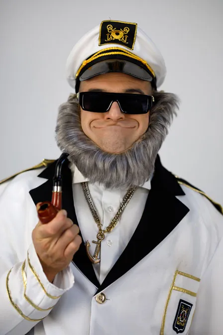 Alter Kapitän Sören Bleibart hat eine Pfeife in der Hand, guckt mit zerknautschtem Gesicht lächelnd in die Kamera, trägt eine weiße Kapitäns-Uniform und schwarze Sonnenbrille, eine Goldkette mit großem goldenen Anker um den Hals
