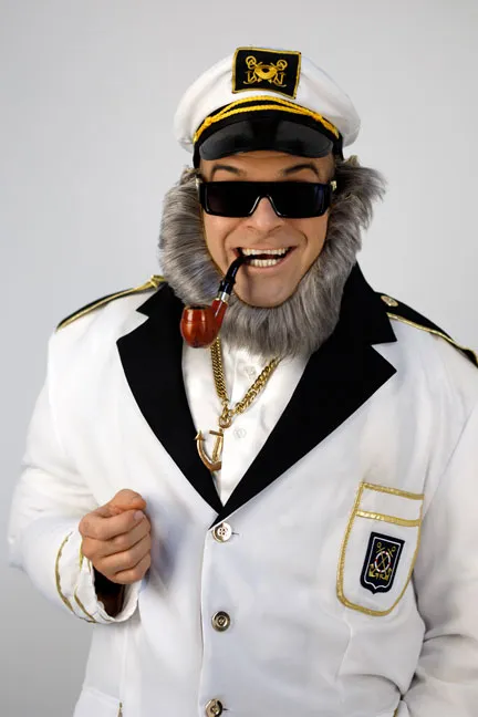 Alter Kapitän Sören Bleibart hat eine Pfeife im Mund, trägt eine weiße Kapitäns-Uniform und schwarze Sonnenbrille, eine Goldkette mit großem goldenen Anker um den Hals