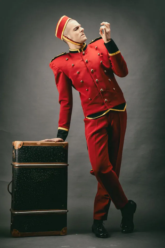 Page Jacques Liqueur trägt eine edle rote Uniform mit goldenen Knöpfen und schwarze Lackschuhe, steht an einem großen schwarzen Koffer und kontrolliert seine Fingernägel