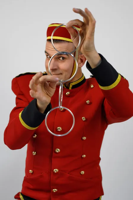 Page Jacques Liqueur trägt eine edle rote Uniform mit goldenen Knöpfen, hält silberne Zauberringe vor seinem Gesicht und guckt durch einen Ring mit seinem Auge in die Kamera